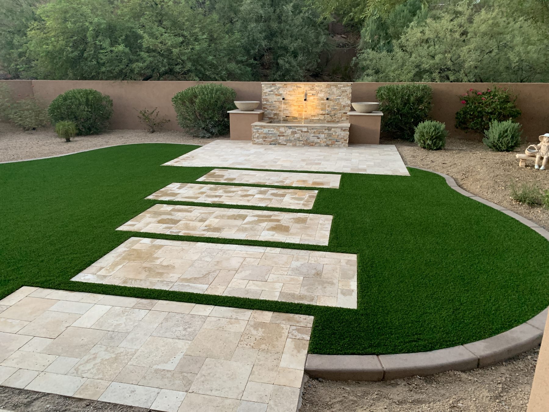 Stone water-feature wall with scupper bowls, turf, and travertine pads — Standard Pools project